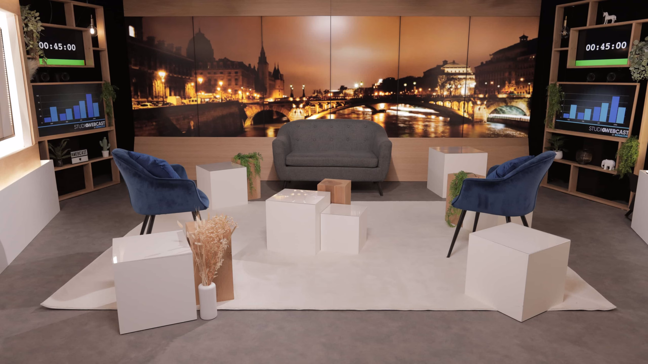 Plateau TV du Studio Webcast en configuration talk show avec canapé, fauteuils velours bleu, carrés blancs et fond Paris by night
