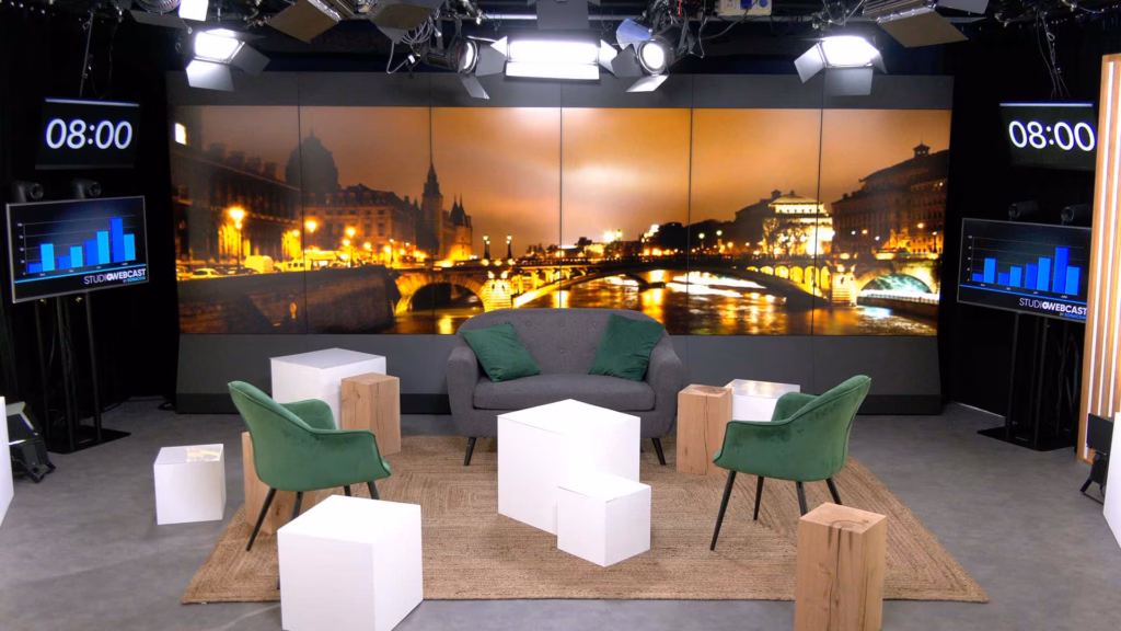 Plateau TV du Studio Webcast en configuration talk show avec fauteuils velours vert, canapé gris, poufs et cubes blancs sur fond Paris by night