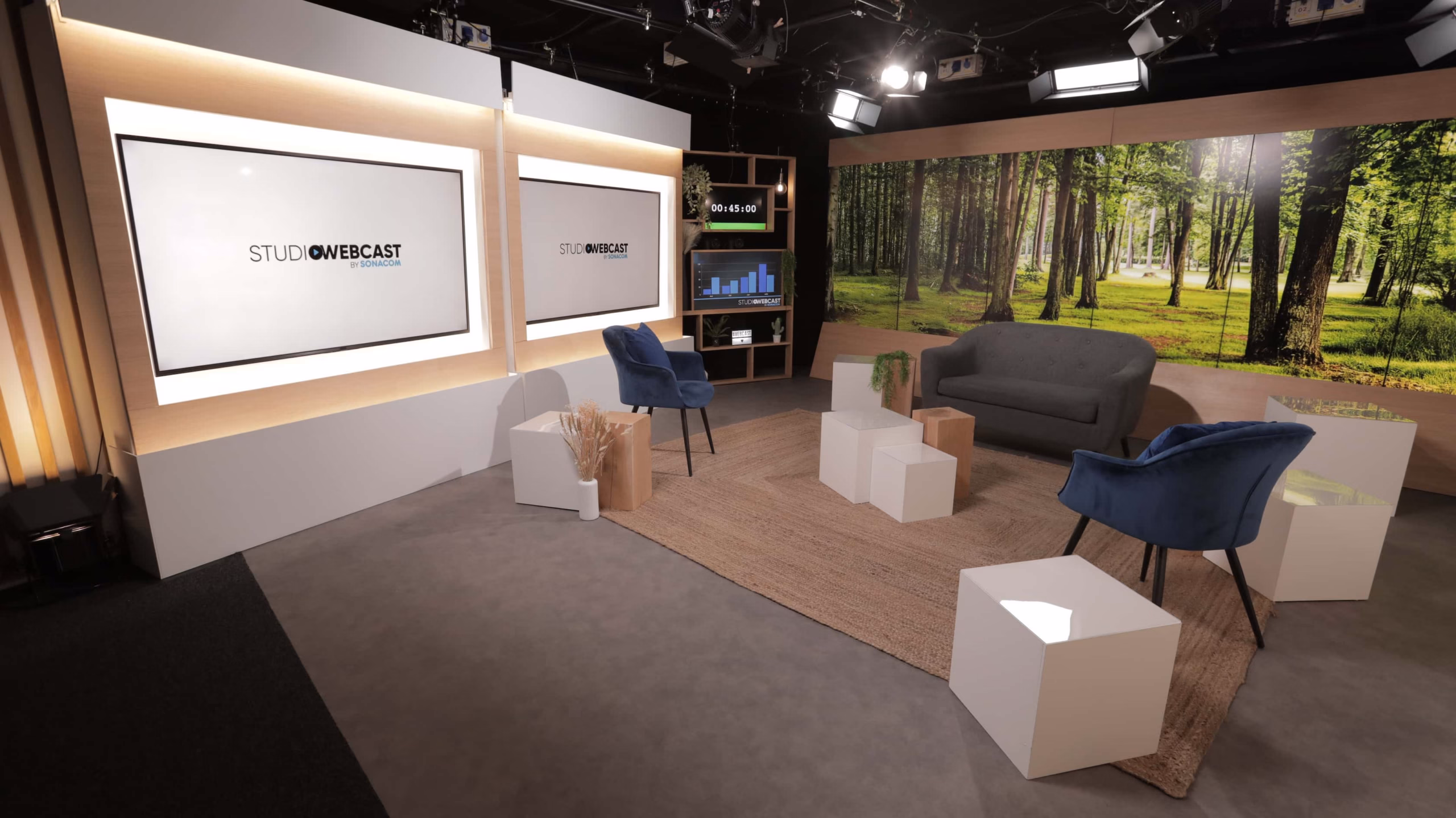 Vue d'ensemble du plateau TV du Studio Webcast en configuration talk show avec canapé, fauteuils velours bleu, cubes blancs et fond LED forêt